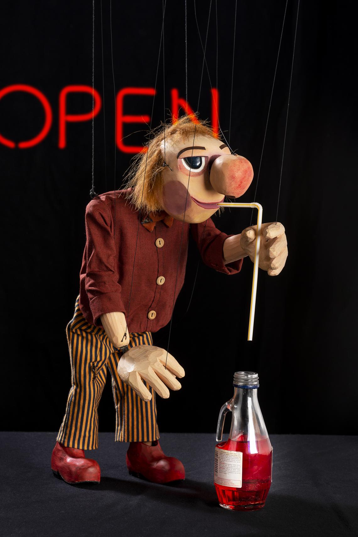 Eine Clowns-Marionette mit einer Art Schweinenase trinkt ein rotes Getränk durch einen Strohhalm.