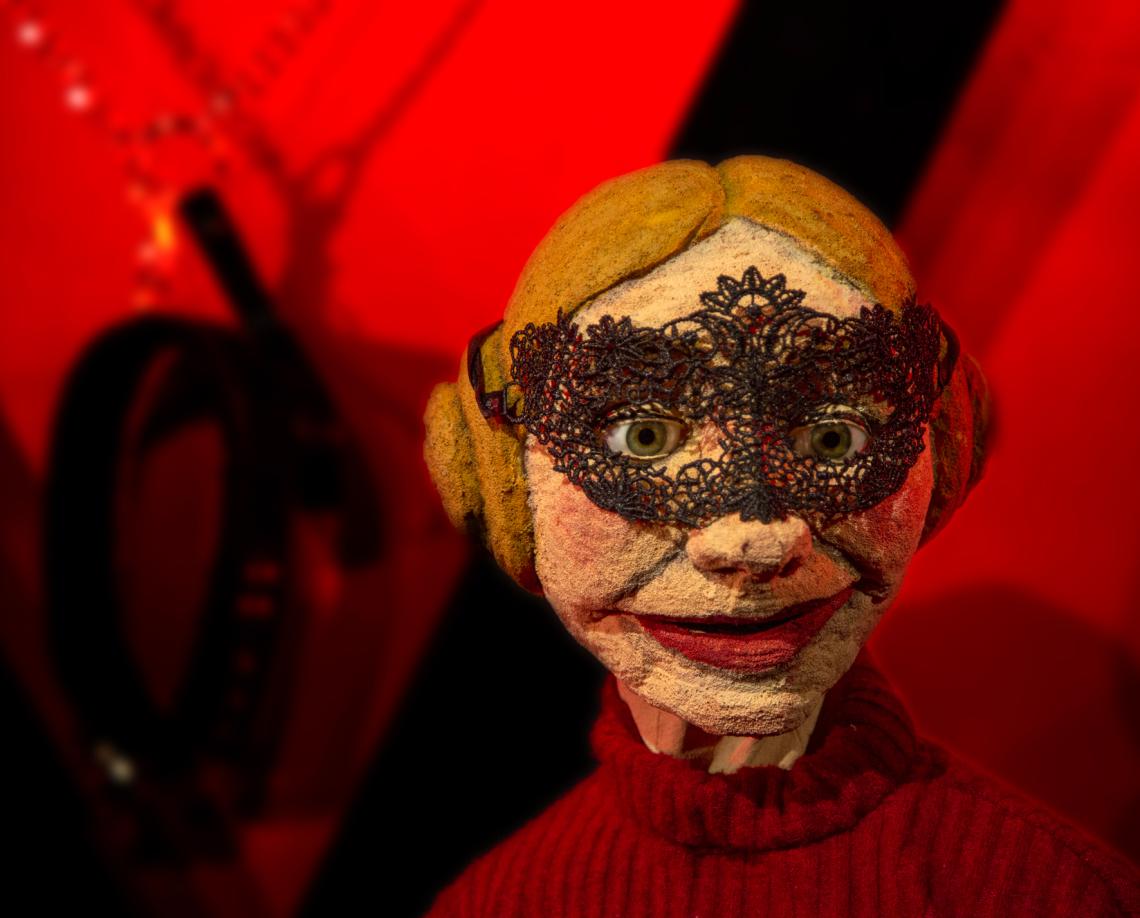 Eine blonde Frau mit schwarzer Halbmaske vor einem roten Hintergrund.