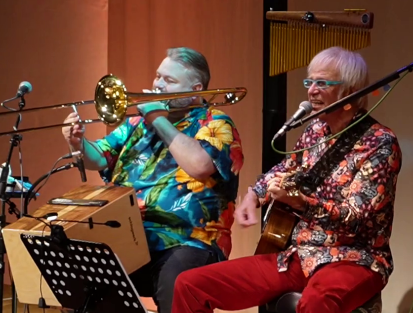 Zwei Musiker in bunten Hemden spielen auf einer Bühne Posaune.