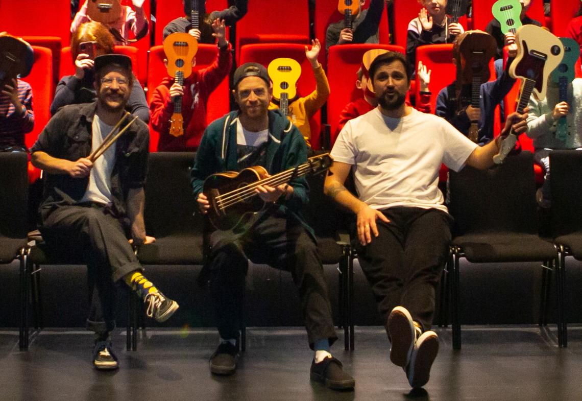 Drei Musiker sitzen mit Ukulelen in der ersten Reihe eines Theatersaals.