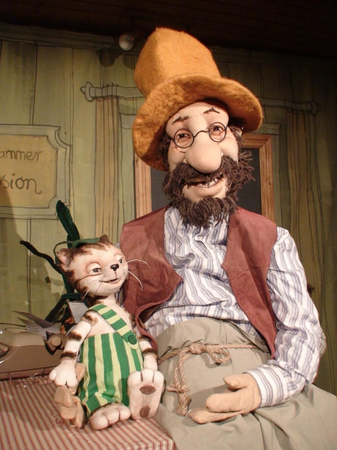 Eine Katze in grüngestreifter Lazhose sitzt auf der Hand eines alten Mannes mit Vollbart, Brille und Hut.