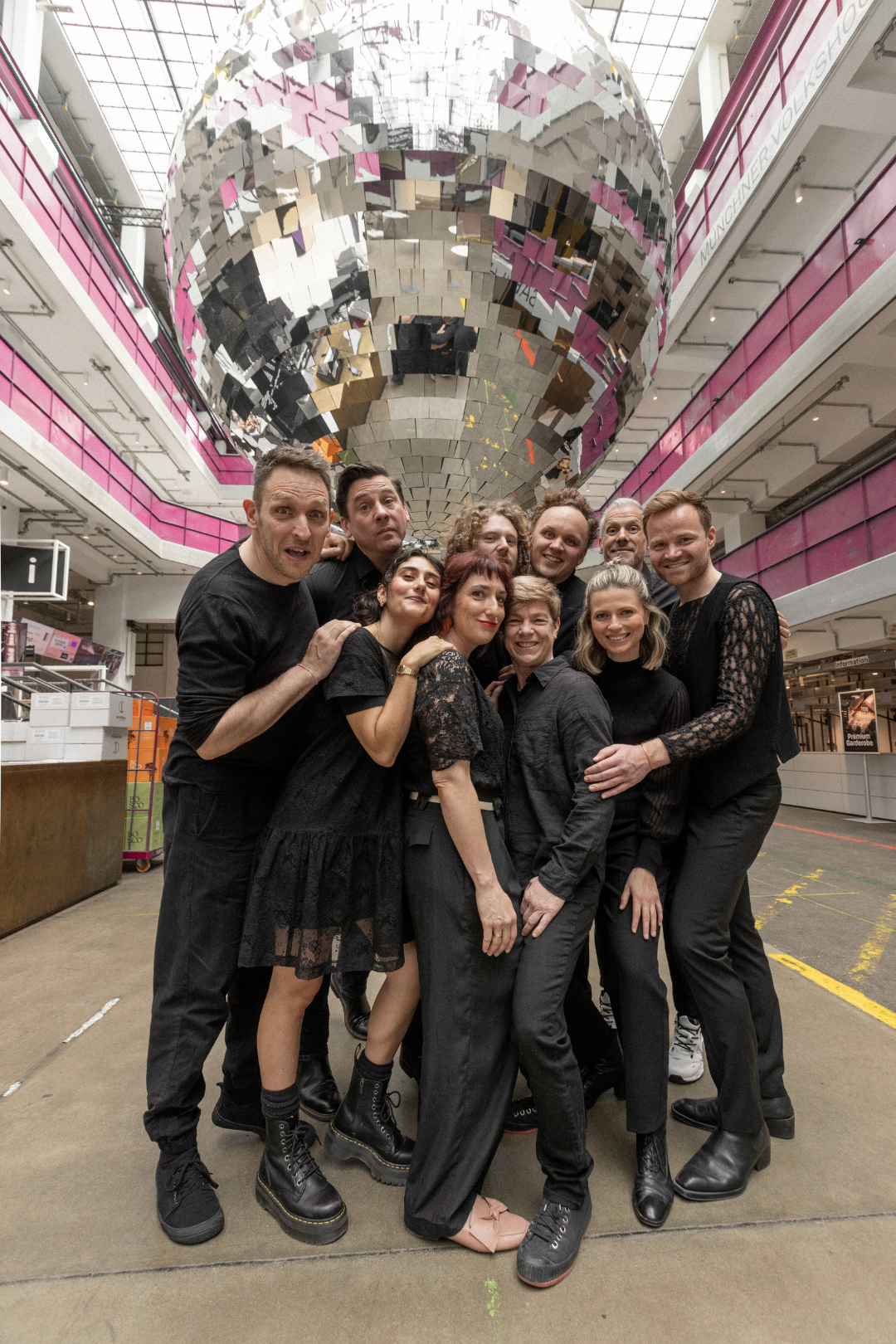 Eine Gruppe von Schauspielenden steht vor einer spiegelnden Kulisse.