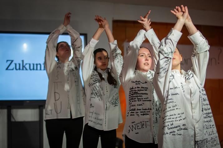 Vier junge Menschen in weißen Kleidern mit Text darauf führen eine Choreografie auf vor einem Bühnenbild, auf dem das Wort "Zukunft" steht.