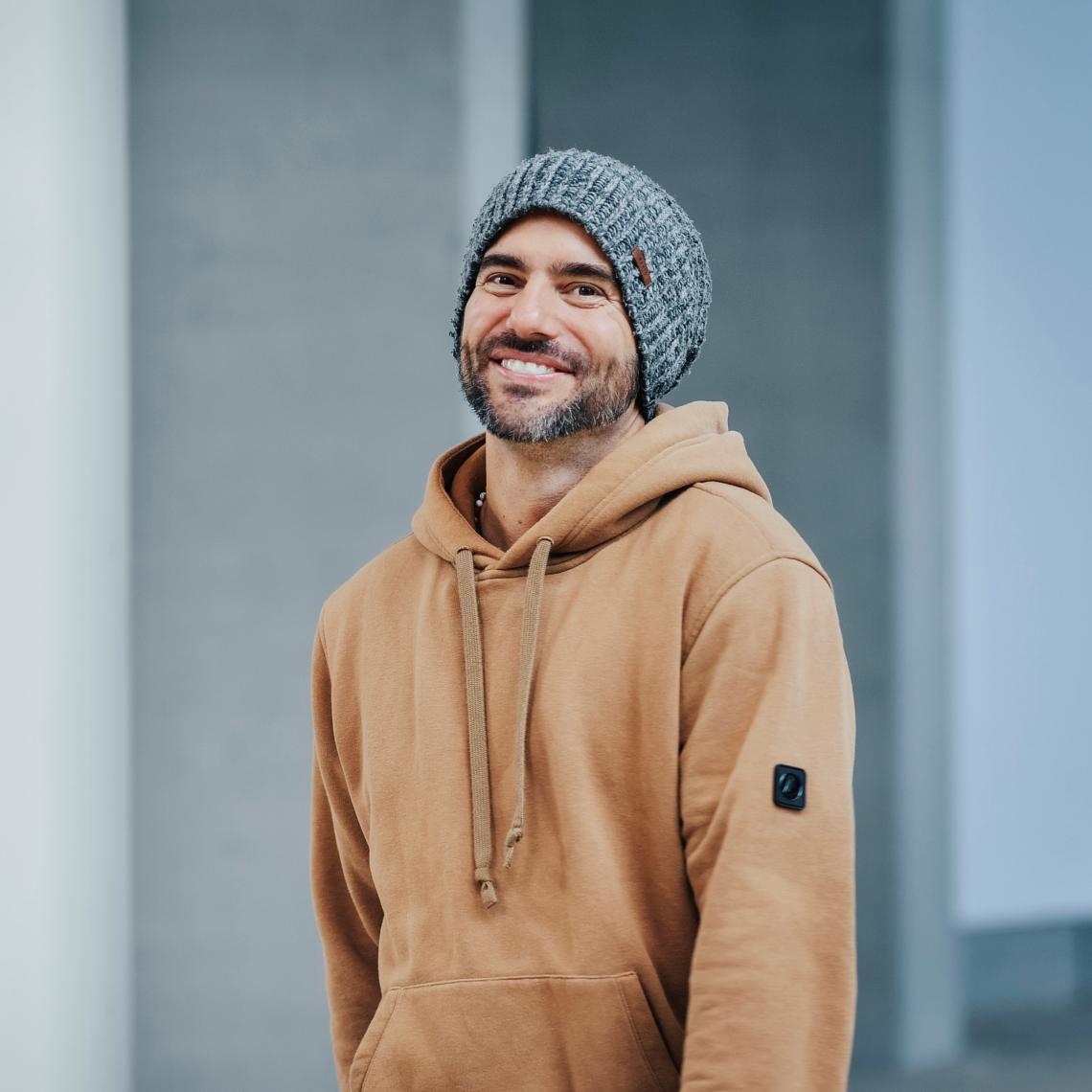 Ein bärtiger Mann mit hellbraunem Kapuzenpulli und grauer Strickmütze lächelt.