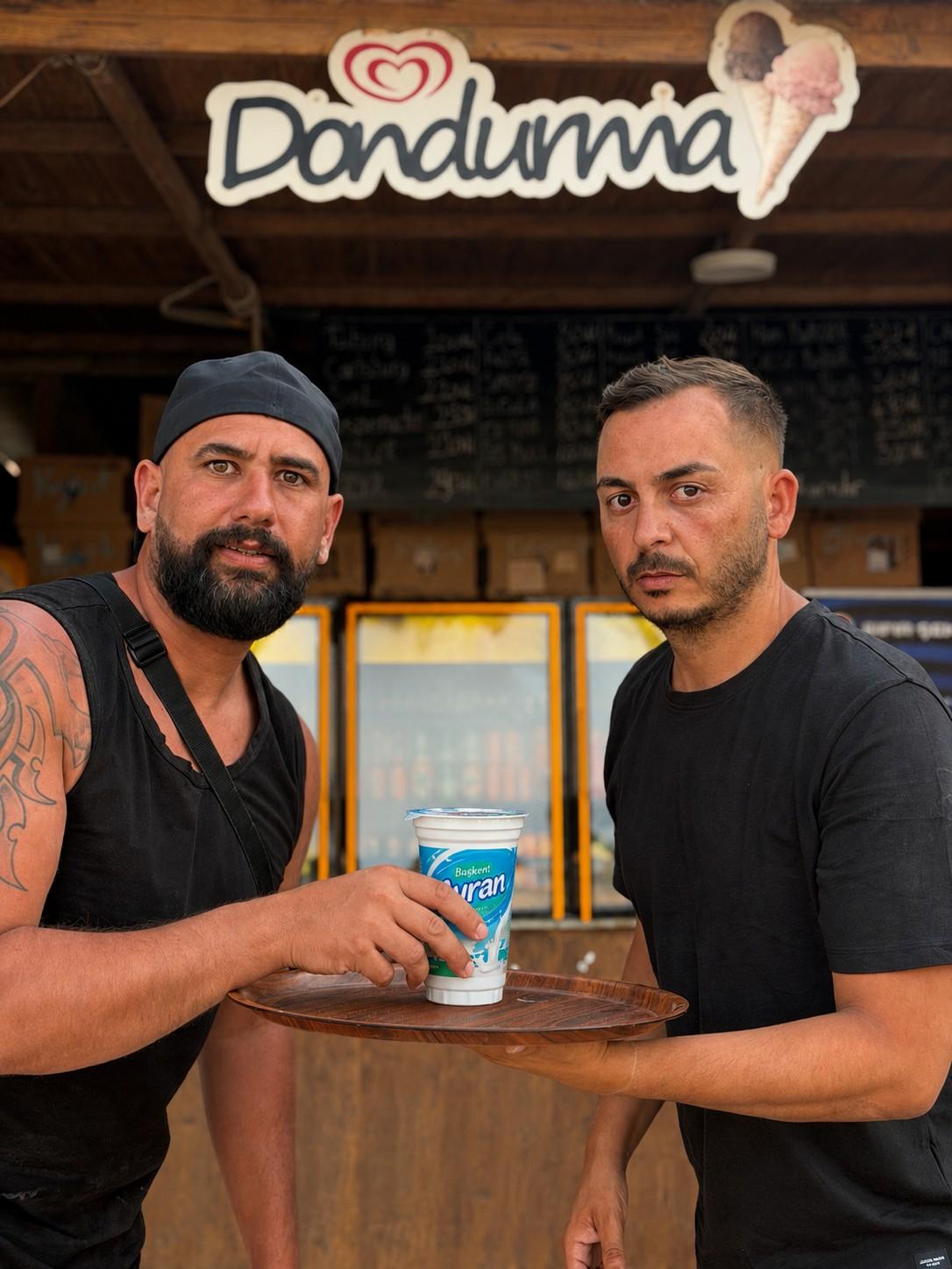 Zwei Männer stehen vor einer Imbissbude mit Eiscreme-Werbung und trinken Ayran.