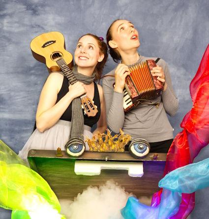 Zwei junge Frauen mit Musikinstrumenten und einem Koffer mit Augen, aus dem Farben sprühen.