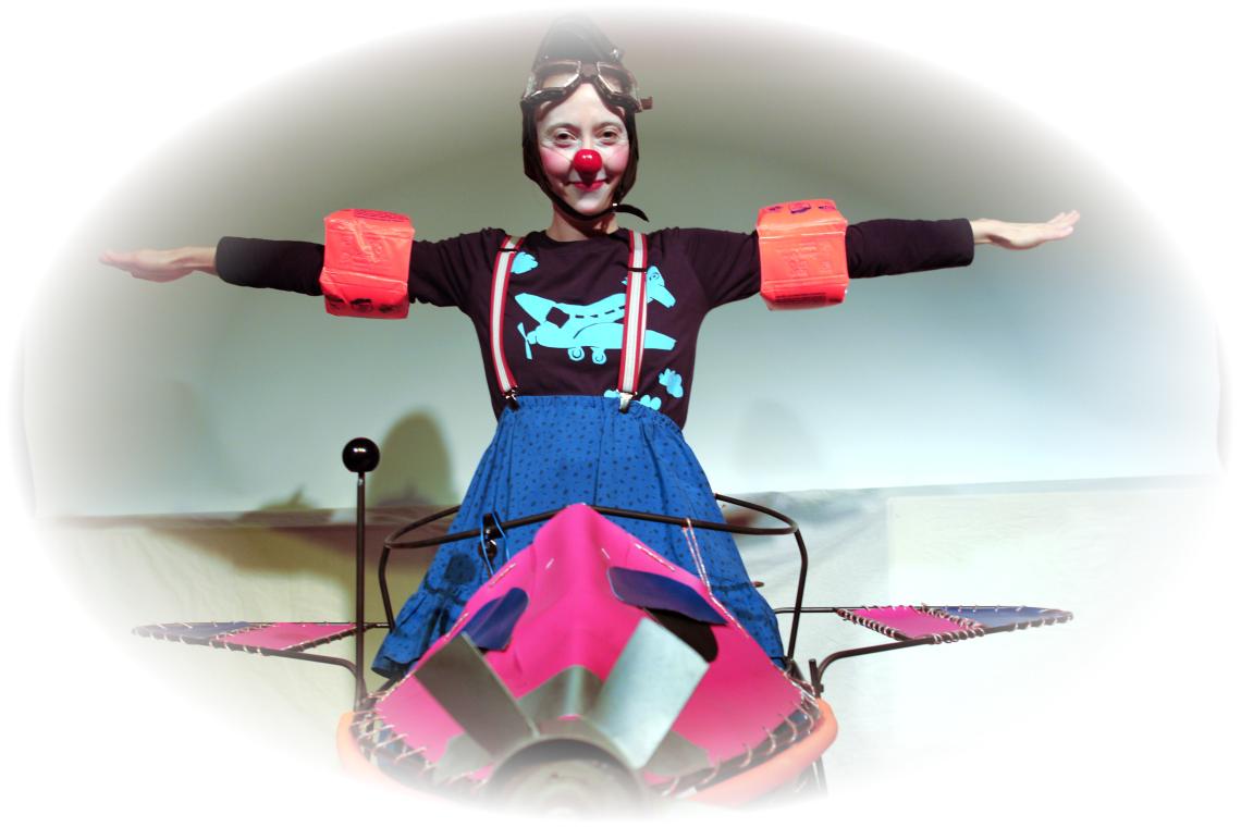 Ein Clownfrau mit roter Nase sitzt in einem Flugzeug und breitet die Arme aus.