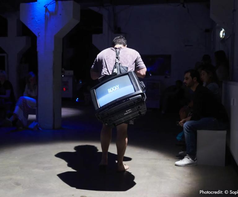 Ein Mann trägt einen laufenden Fernseher auf dem Rücken.