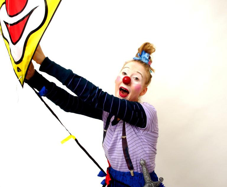Eine junge Frau mit Clownsgesicht hält einen Drachen fest, auf dem ebenfalls ein Clownsgesicht zu sehen ist.