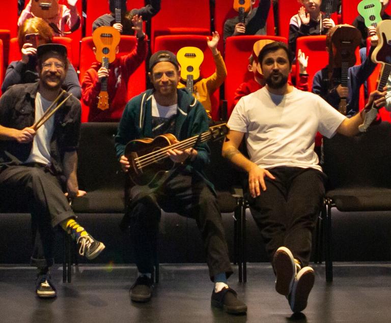 Drei Musiker sitzen mit Ukulelen in der ersten Reihe eines Theatersaals.