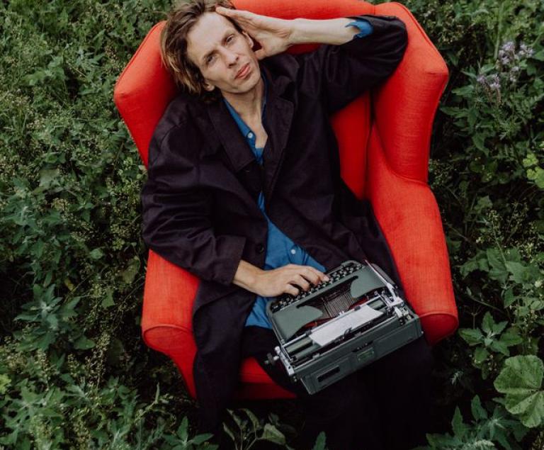 Ein Mann sitzt mit einer Schreibmaschine auf dem Schoß in einem roten Sessel auf einer Wiese.