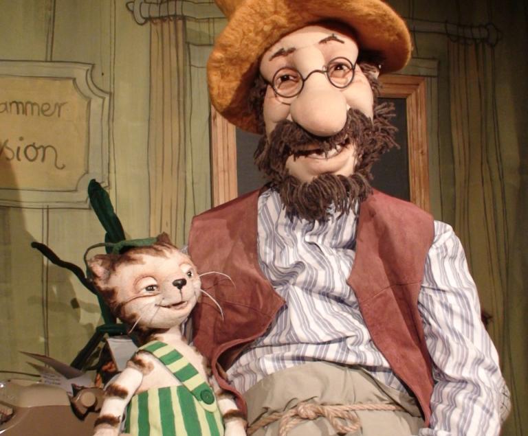 Eine Katze in grüngestreifter Lazhose sitzt auf der Hand eines alten Mannes mit Vollbart, Brille und Hut.