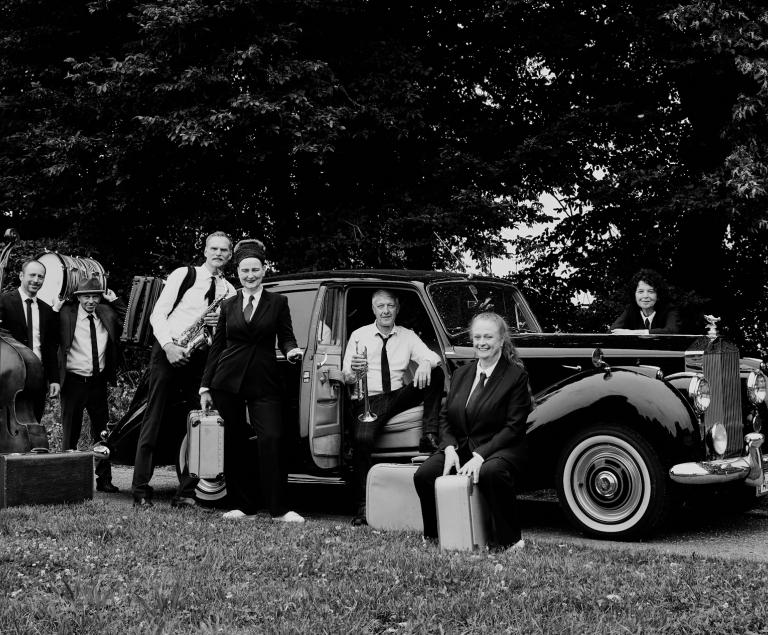 Eine Swingband in amerikanischer Kleidung der 40er-Jahre posiert vor einem Oldtimer.