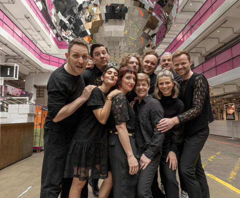 Eine Gruppe von Schauspielenden steht vor einer spiegelnden Kulisse.