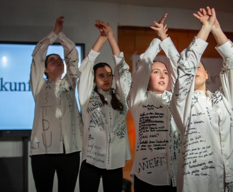 Vier junge Menschen in weißen Kleidern mit Text darauf führen eine Choreografie auf vor einem Bühnenbild, auf dem das Wort "Zukunft" steht.