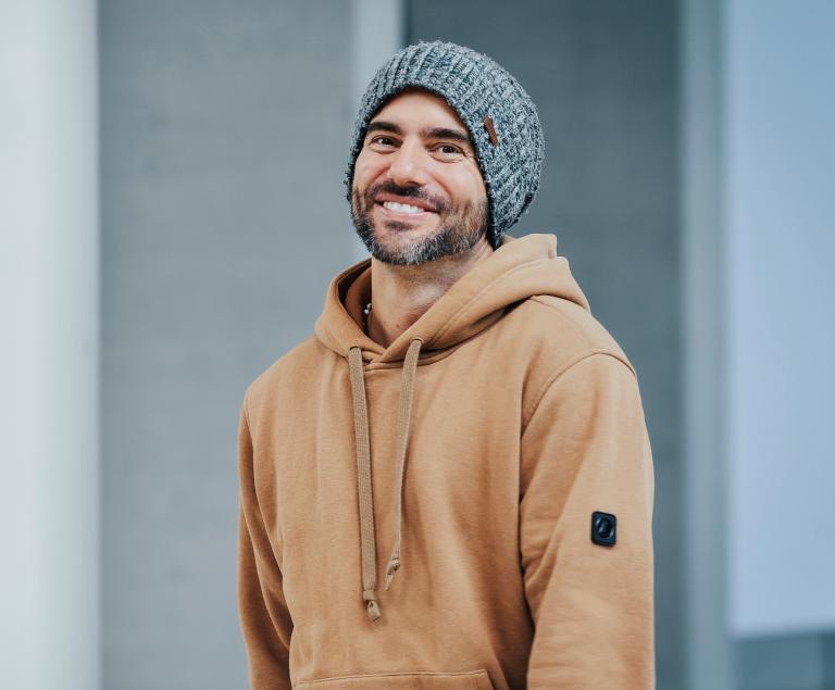 Ein bärtiger Mann mit hellbraunem Kapuzenpulli und grauer Strickmütze lächelt.