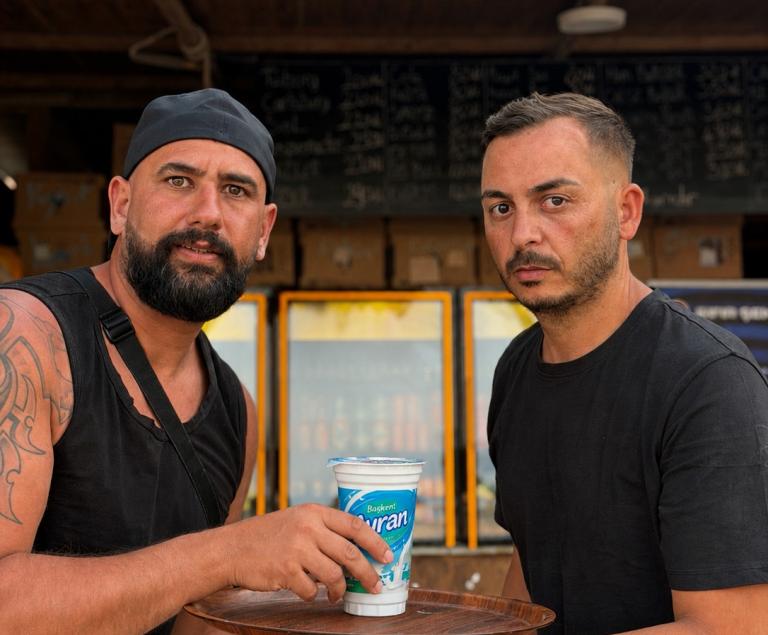 Zwei Männer stehen vor einer Imbissbude mit Eiscreme-Werbung und trinken Ayran.