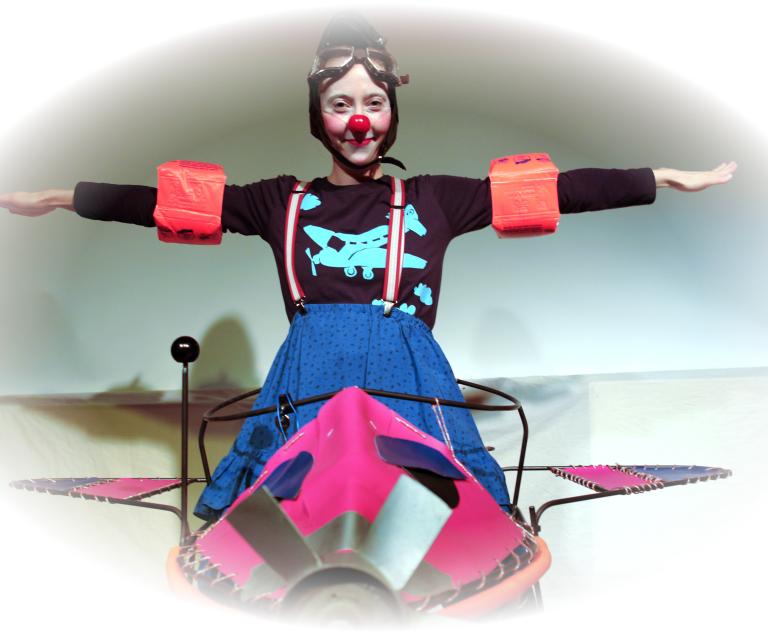 Ein Clownfrau mit roter Nase sitzt in einem Flugzeug und breitet die Arme aus.